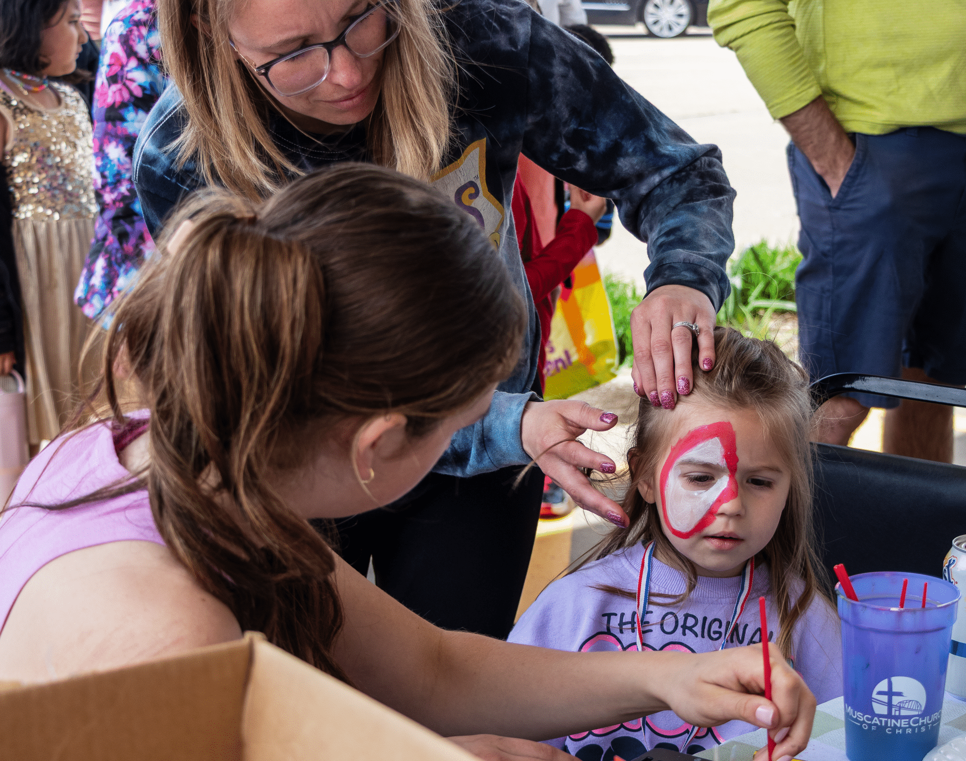 Two women lovingly paint a child’s face at our joyful outdoor gathering, creating smiles and memories together. - Muscatine Church of Christ