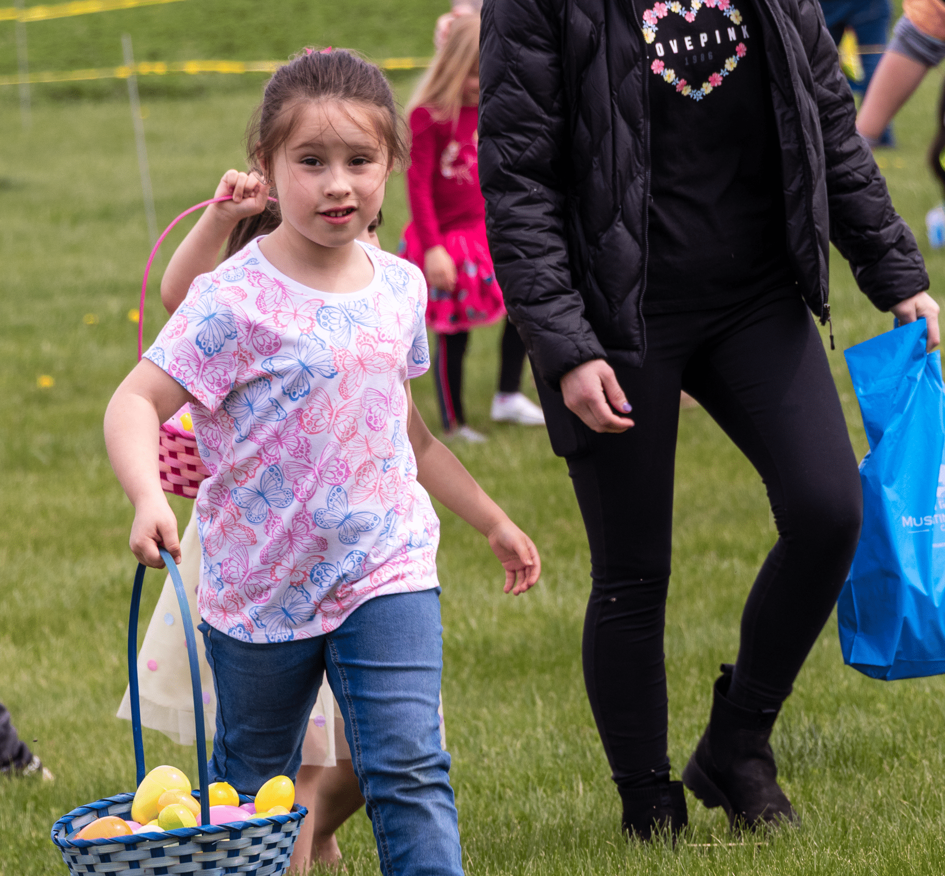 A young girl with a blue basket joins smiling friends and family on the grass, sharing joyful moments together. - Muscatine Church of Christ