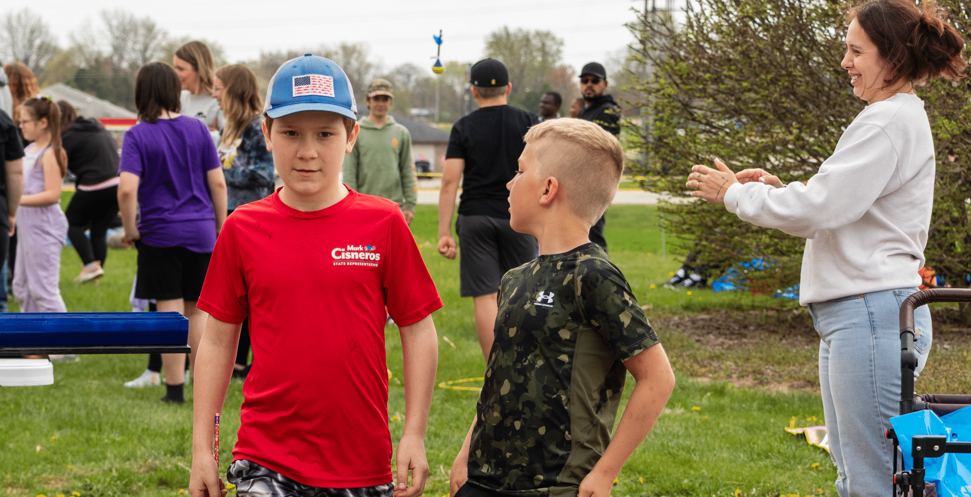 Two boys smile together on the grass as friends cheer nearby—everyone is welcome in our joyful community. - Muscatine Church of Christ