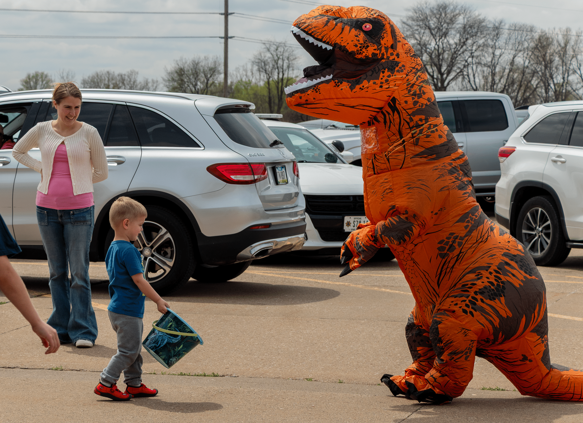 A friendly dino greets a child with a basket, sharing joy and welcome in our church family parking lot. - Muscatine Church of Christ