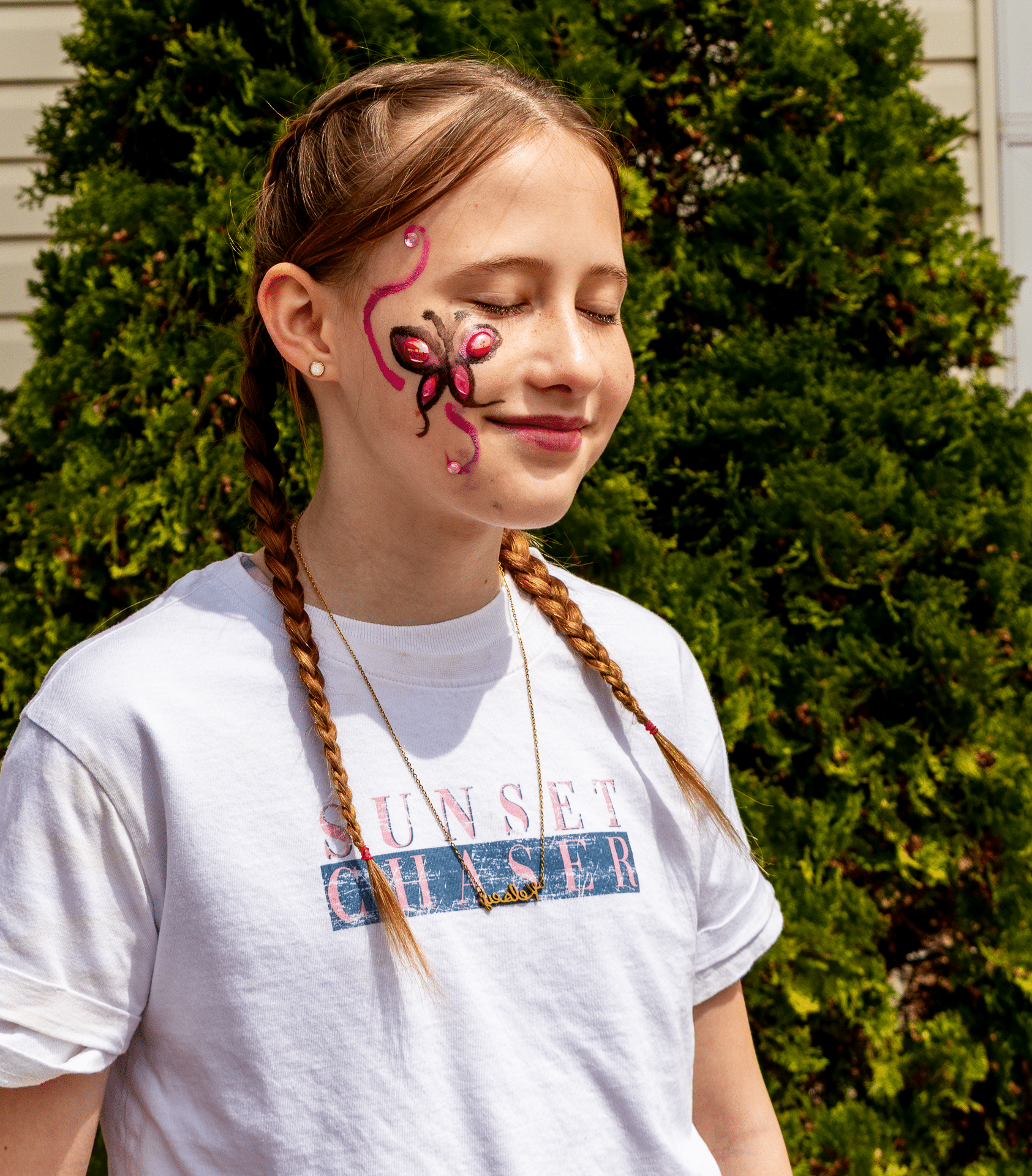 Young girl with braided hair and butterfly face paint, eyes closed in peaceful joy, stands outside by green bushes. - Muscatine Church of Christ